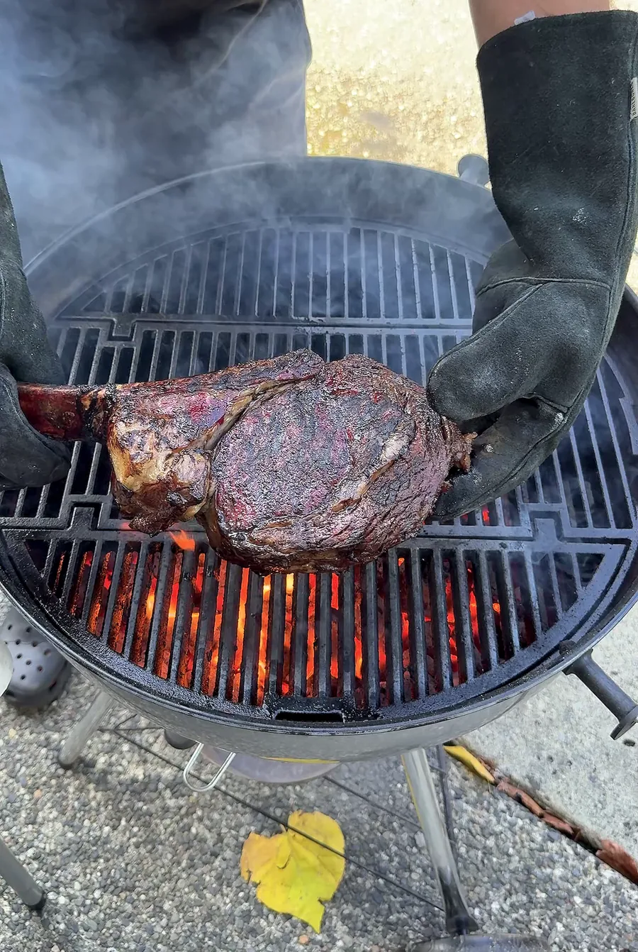 Gloved hands holding the seared tomahawk above the kettle fire